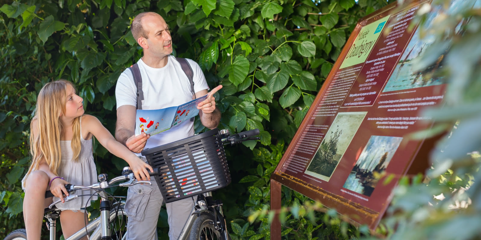 Photo d'un père et sa fille à vélo se renseignant sur un panneau du musée de Daubigny à Auvers-sur-Oise